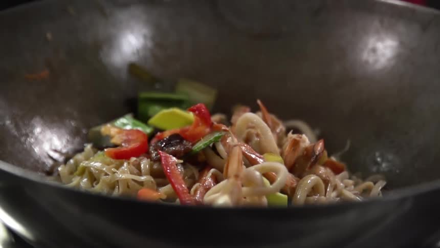 Shrimp and cut vegetables are dropped atop a wok filled with noodles and other vegetables
