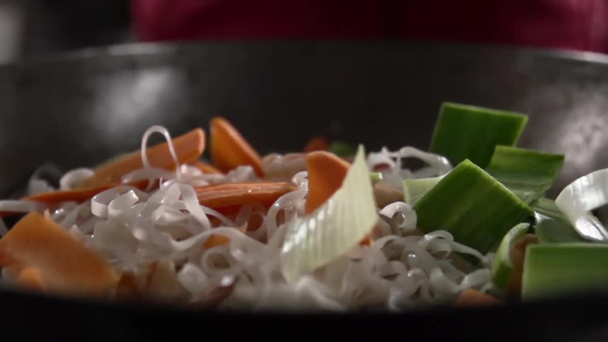 Chopped vegetables falling on top of a wok filled with noodles and shrimps