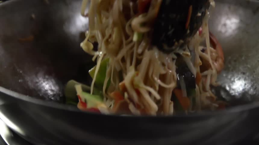 Noodles and vegetables being tossed in a wok with chopsticks and a spoon