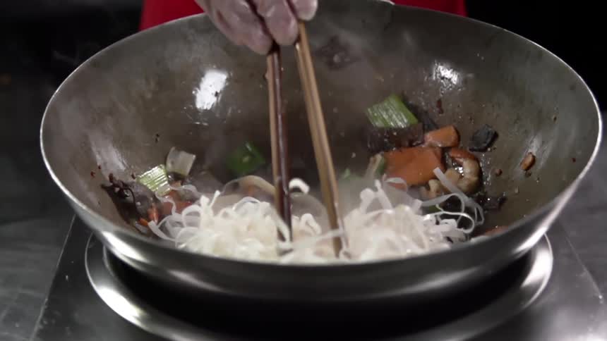 A chef tilts and swirls a wok pan as he stirs noodles shrimp and cooked vegetables with chopsticks