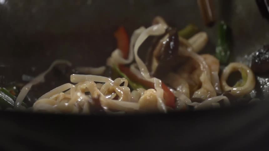 A person stirs noodles shrimp and vegetables together in an oily wok