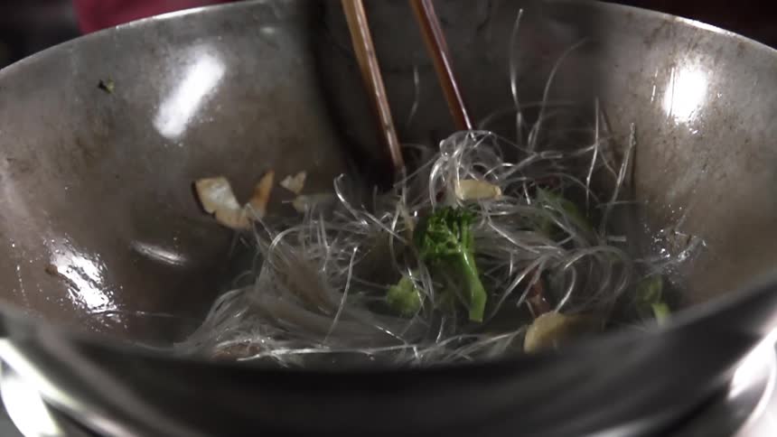 A pair of chopsticks is used to stir together vegetables and rice noodles in a glass bowl