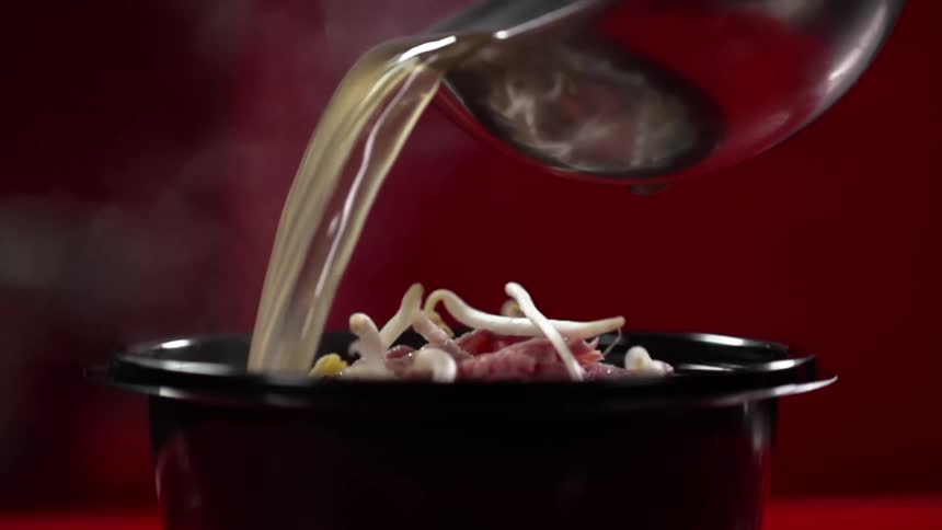 Broth is poured from a ladle into a black ceramic bowl filled with sliced pork and bean sprouts