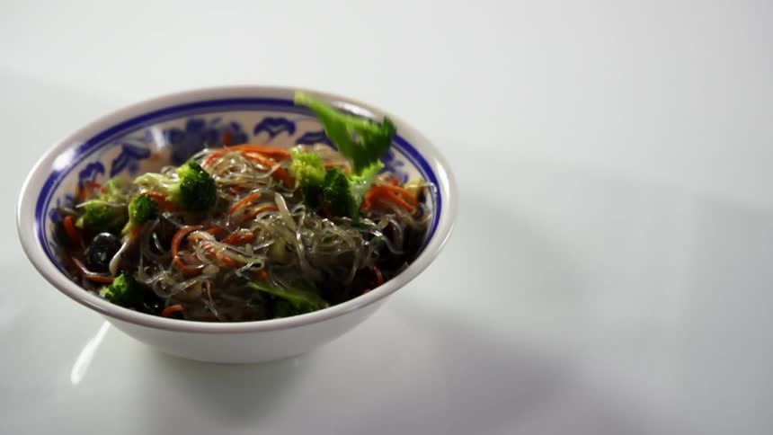 Broccoli falling into a bowl filled with glass noodles on a white table
