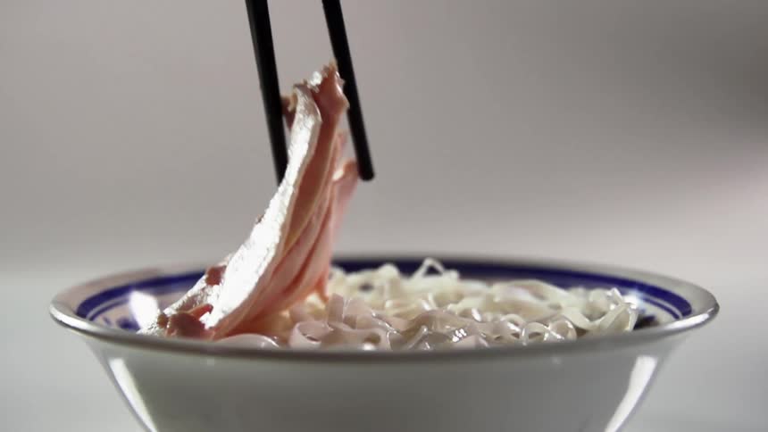 Slice of cooked meat placed over a bowl of noodles using chopsticks