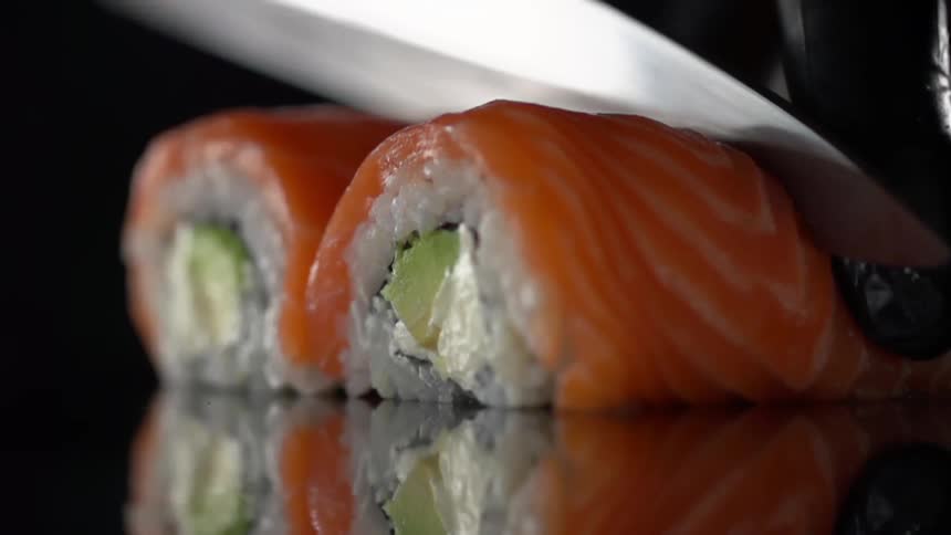 A sharp knife slowly cuts through a sushi roll made with cucumber wrapped with rice and raw fish
