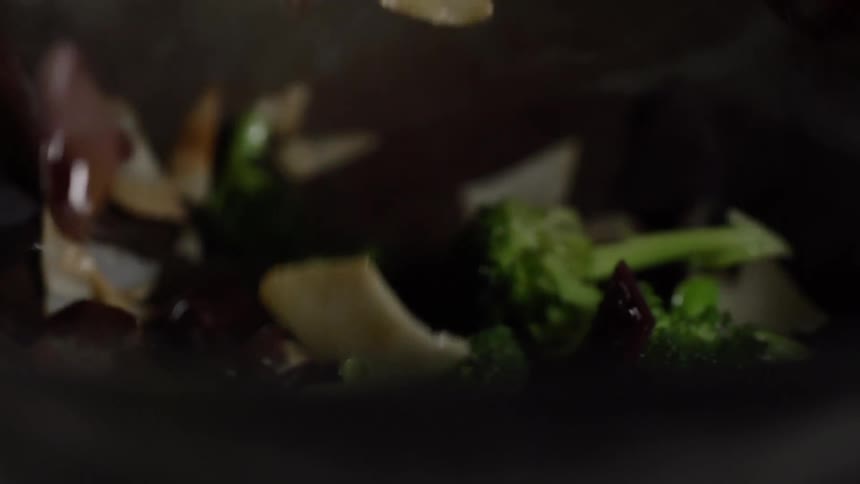 Large wooden sticks and slotted utensil stir vegetables in wok