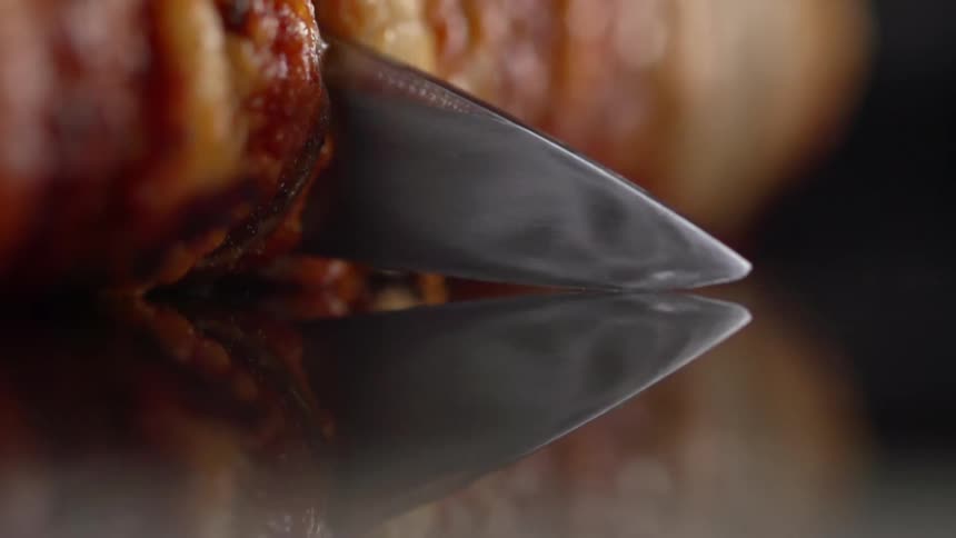 Sharp kitchen knife cuts a roll of meat over a reflective surface