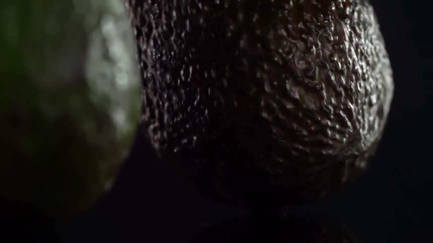 Avocados drop on a surface bouncing and rolling away against a black background
