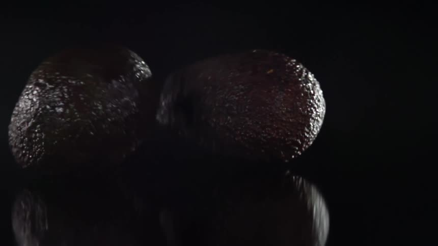 Avocados drop onto flat reflective surface in dark room