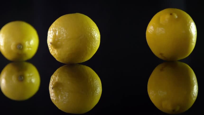 Three fresh lemons are dropped onto a dark and highly reflective surface where they gently bounce and roll around