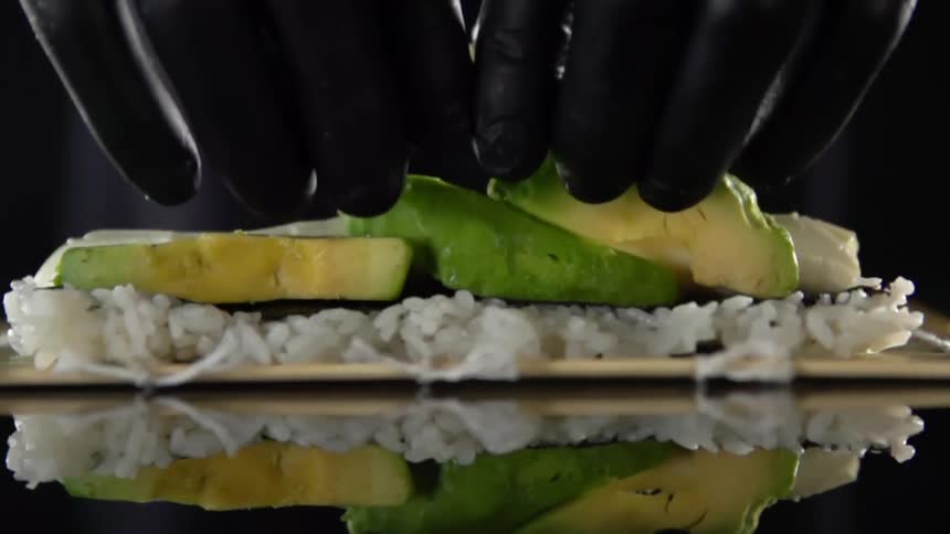 A person wearing black disposable gloves carefully places several pieces of sliced avocado atop a pile of white rice