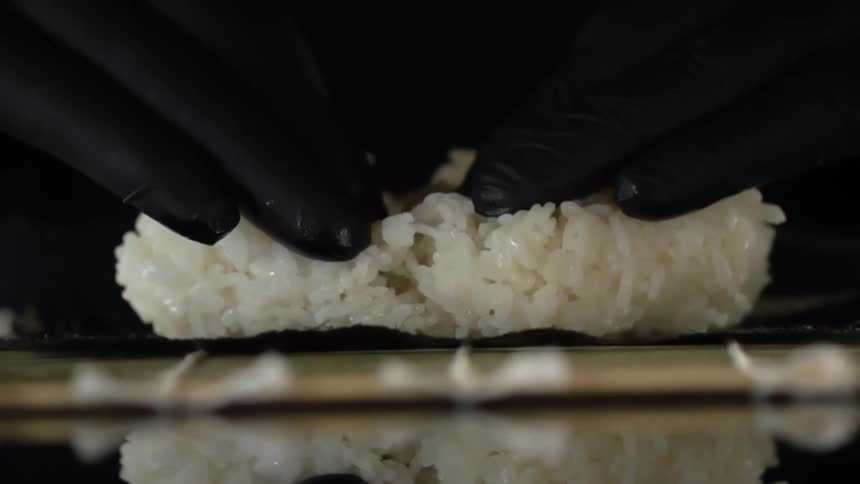 Two hands covered with black latex gloves spread a ball of white sticky rice across a piece of nori to make sushi