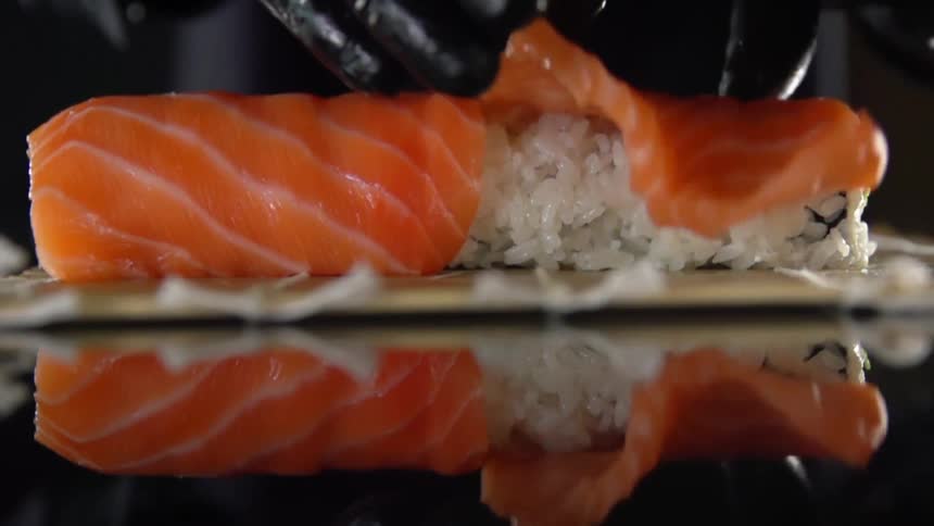 A fillet of raw fish being laid atop a block of rice on a sushi rolling mat