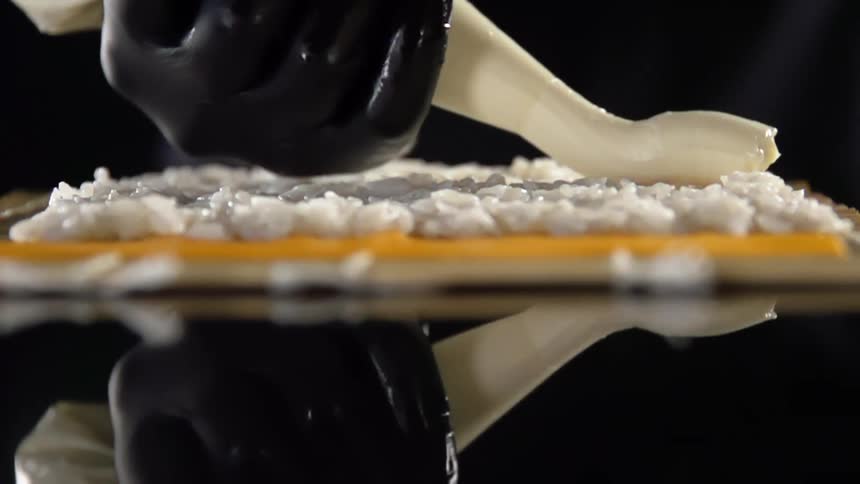 A person wearing black disposable gloves carefully applies a layer of paste atop a sheet of cooked rice