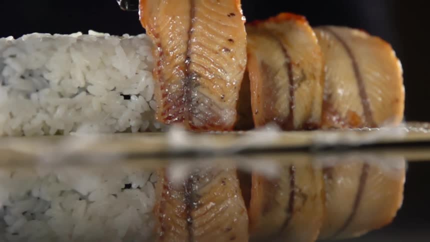 Chef with black latex gloves places fish fillets around a roll of sushi over a bamboo mat