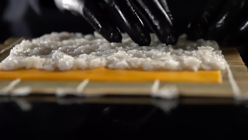 A person wearing black plastic gloves picks and spreads rice across a sushi rolling mat