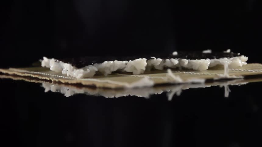 Chef wearing black latex gloves places rice on seaweed over a bamboo mat in a reflective surface