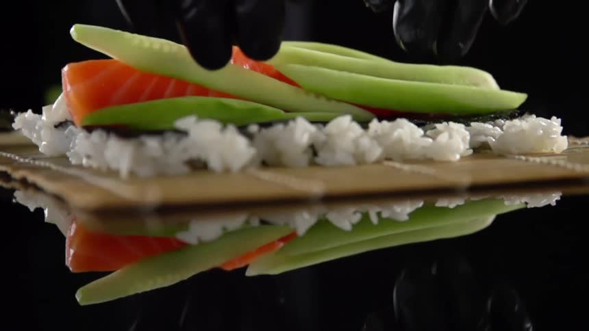 A person wearing black disposable gloves places some fresh green cucumber spears atop raw fish on a pile of rice