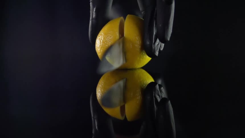 Mirrored reflection on black backdrop of a hand wearing a black glove as a knife slices the lemon in half widthwise