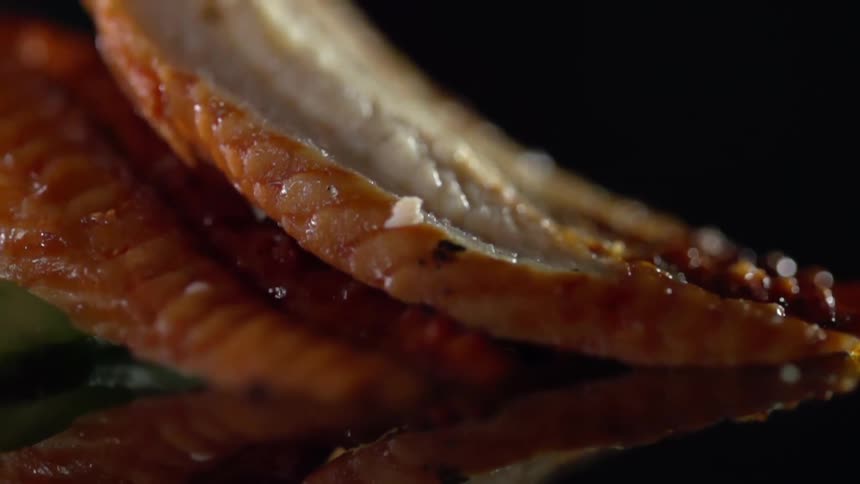 Slices of cooked fish stacked over a reflective surface against a black background