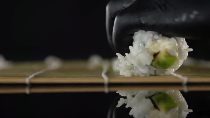 Chef wearing black latex gloves rolls sushi over a bamboo mat