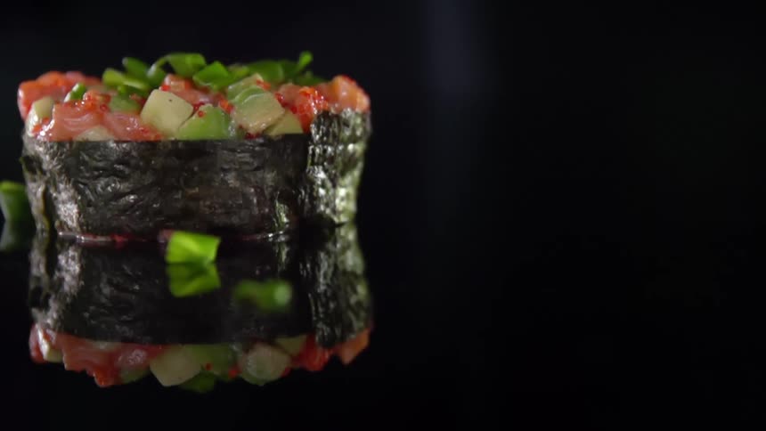 Some final ingredients are dropped onto the top of a sushi roll wrapped in seaweed