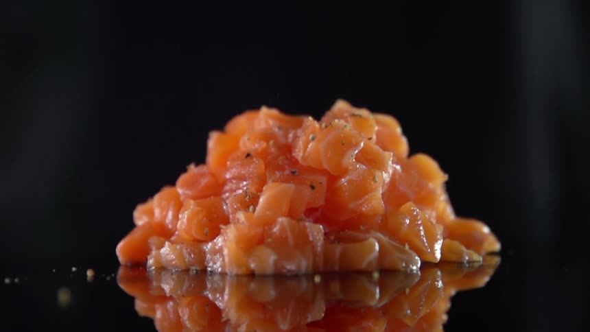 A pile of diced raw fish sits atop a dark reflective surface as seasoning is sprinkled over it