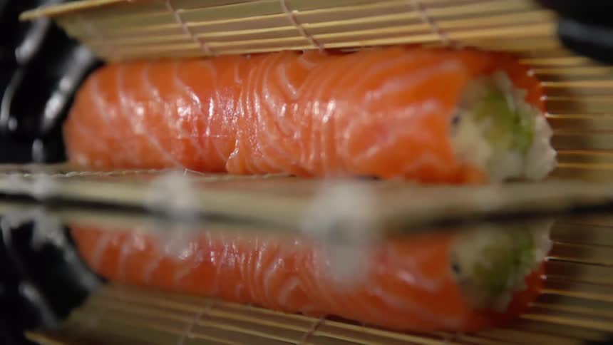Chef with black latex gloves rolls a salmon sushi using a bamboo mat over a reflective surface