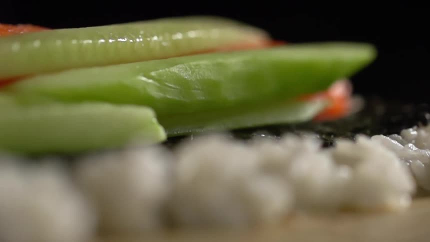 Person wearing black gloves add vegetables to fish on seaweed on bed of rice