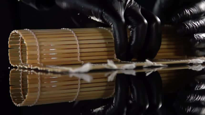 Chef wearing black latex gloves rolls sushi using a bamboo mat over a reflective surface