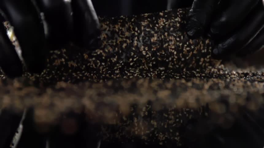 Chef wearing black latex gloves rolls sushi over sesame seeds on a reflecting surface