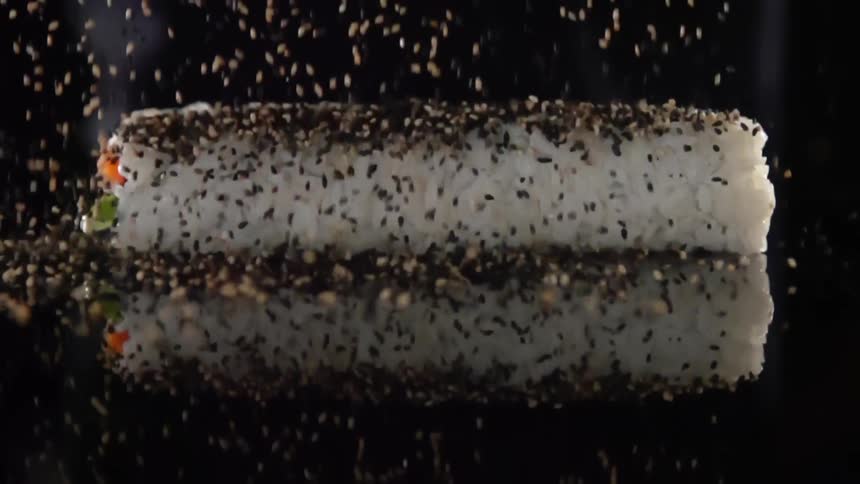 Black sesame seeds rain down on an inside out sushi roll on a mirror surface