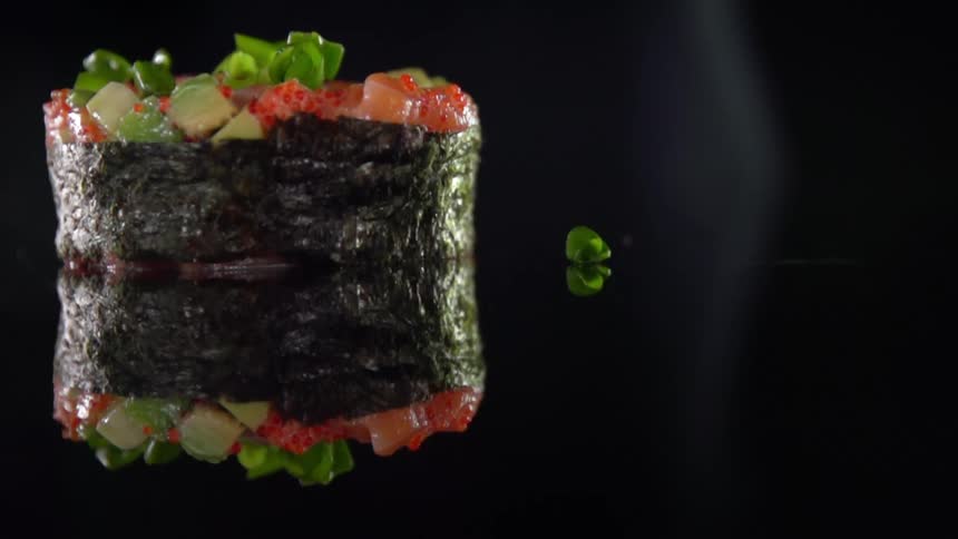 A round piece of sushi wrapped in seaweed sits atop a dark reflective counter while some additional cut vegetables are dropped on top of it
