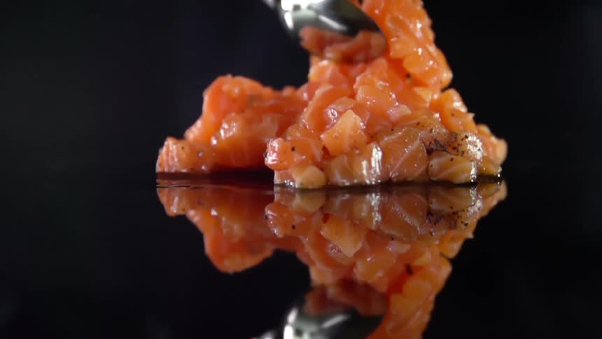 A pile of chopped fish with pepper and seasoning is stirred with a spoon atop a dark highly reflective surface