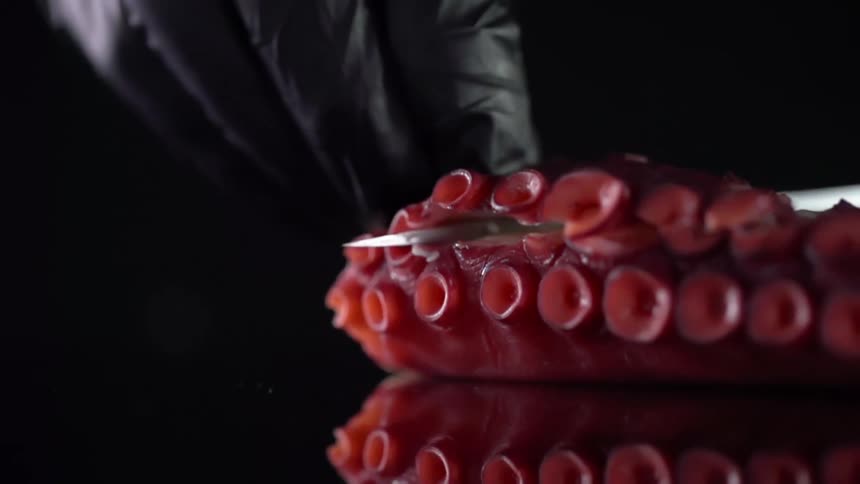 A person with black gloves cutting an octopus with a knife on a glass table