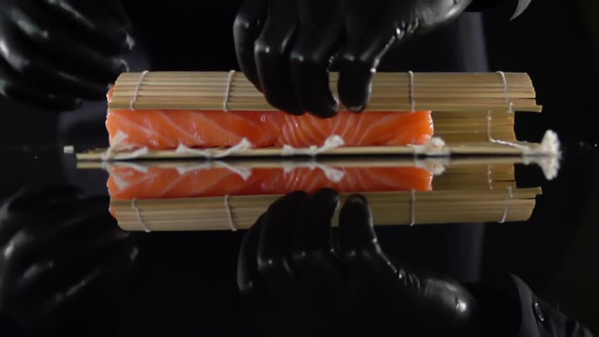 Chef wearing black latex gloves wraps a salmon sushi roll with a bamboo mat