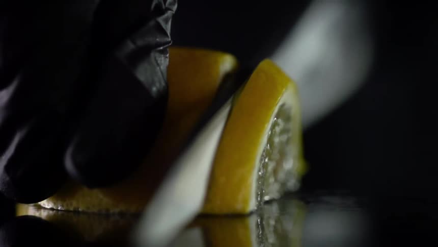 A person slices a lemon with a knife on a dark colored table