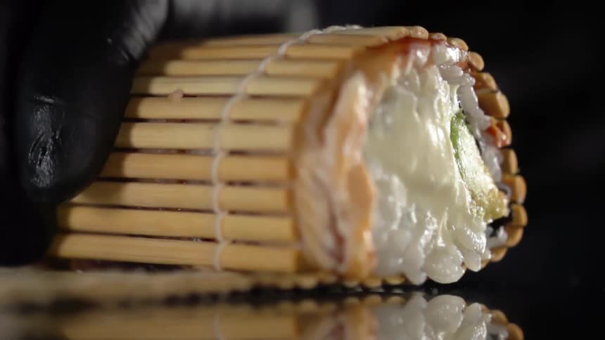 Black gloved hand rolls food inside of a bamboo container