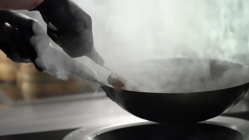 A person holds a steaming hot wok and tosses the food inside to mix it while stirring over heat