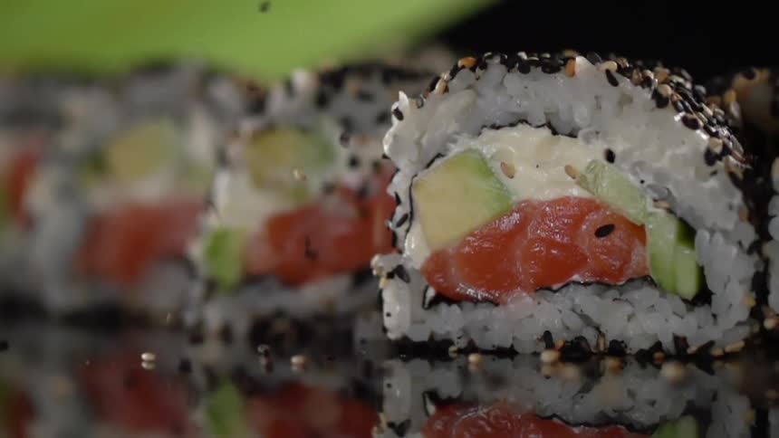 Sesame seeds falling onto a row of sushi rolls on a table