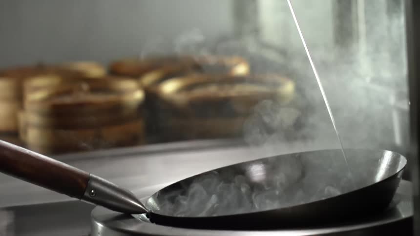 Smoke rises as cooking oil is poured into a hot wok in a kitchen