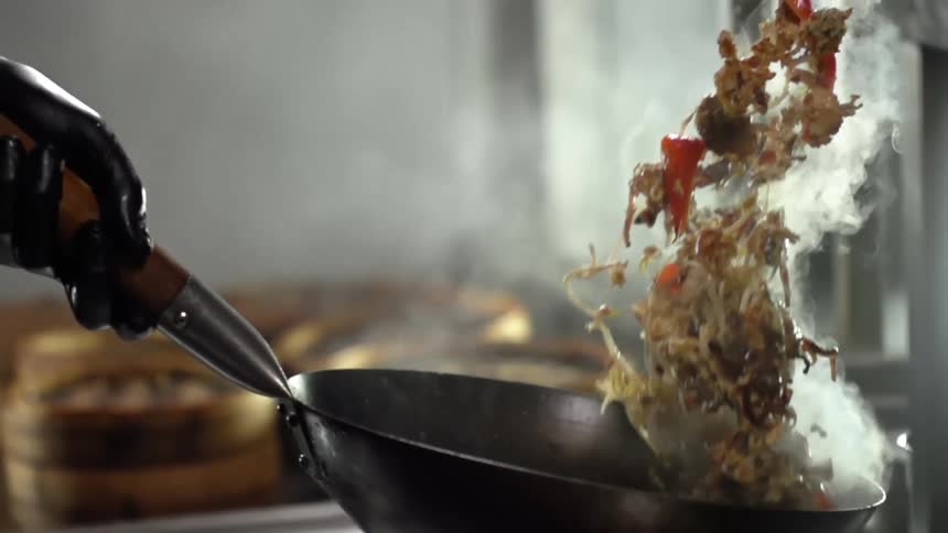 Person wearing black gloves tosses stir fry in wok while cooking near steam baskets