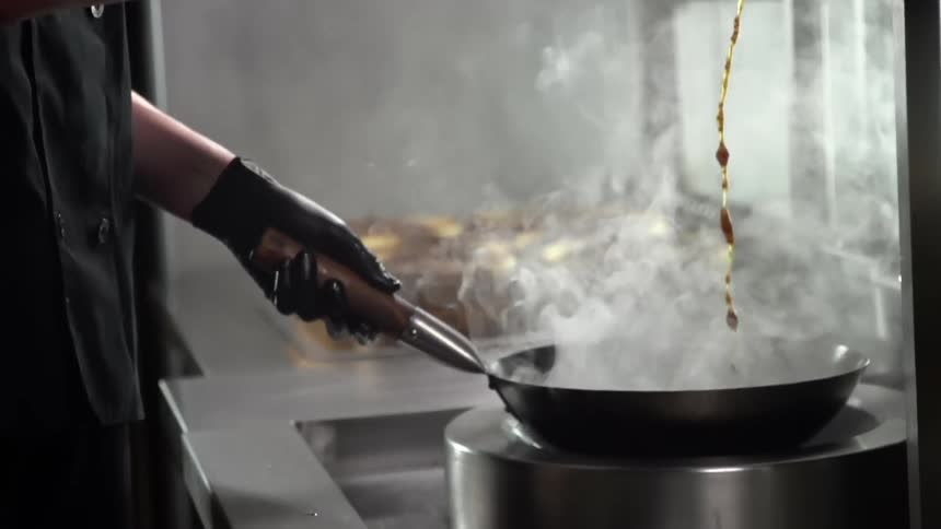 A cook pours a dark sauce from a ladle into a smoking skillet