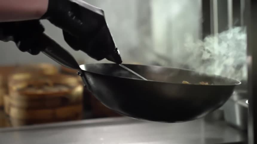 Chef wearing black latex gloves toss food inside a hot steaming wok