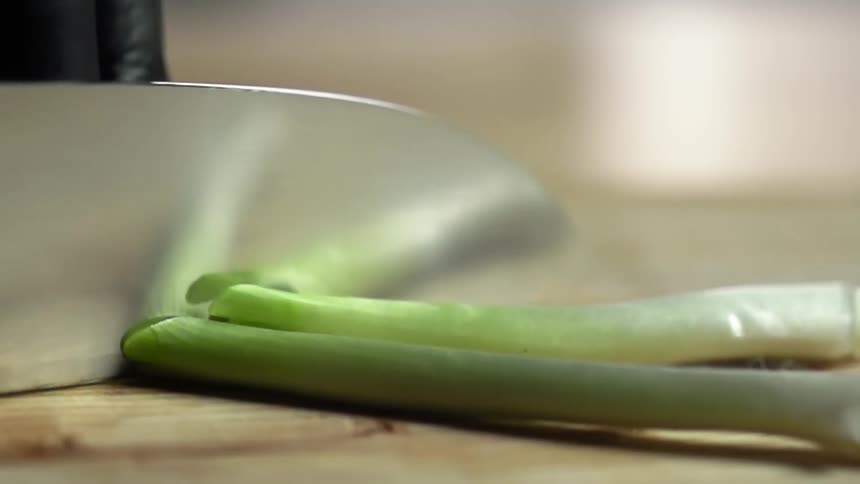A person with black gloves cutting green onions with a knife on a cutting board