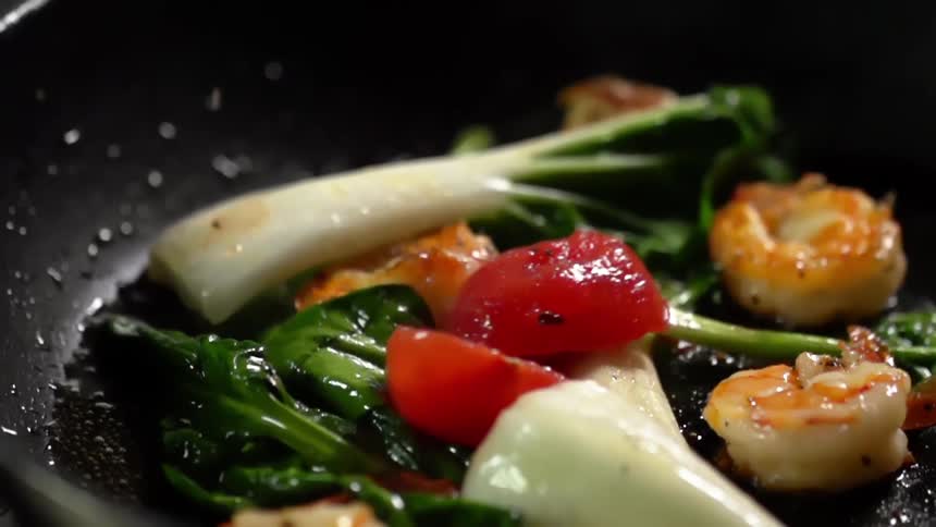 Vegetables and shrimps are being cooked in a black pan