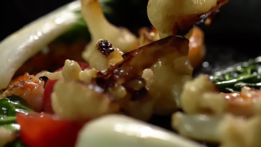 Cooked cauliflower bits fall inside an iron cast pan with vegetables and shrimp