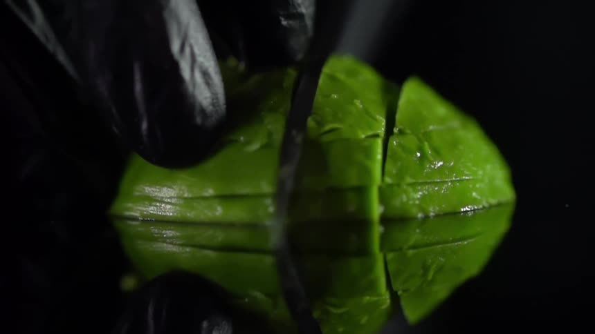 A person wearing black disposable gloves slowly cuts a avocado half into slices with a knife