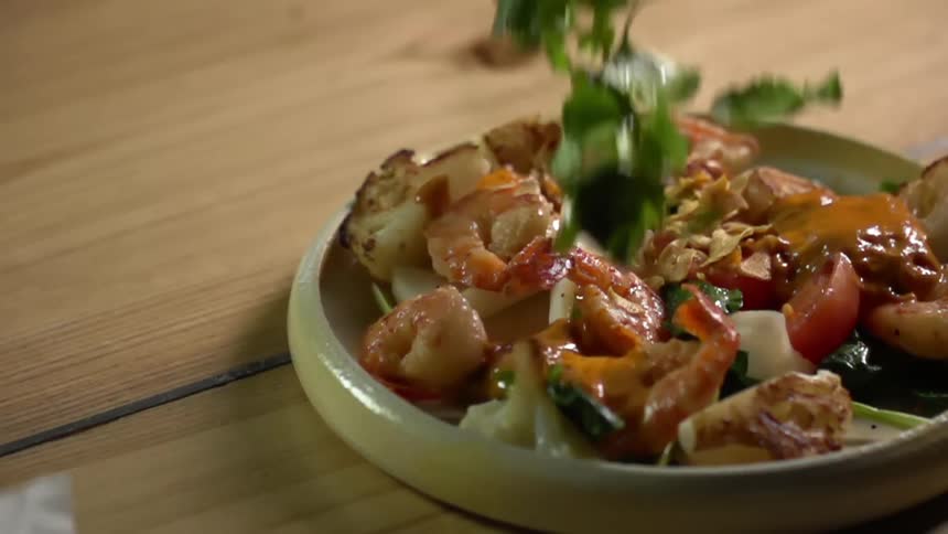 Leafy green vegetables falling into a dish containing cooked shrimps and vegetables